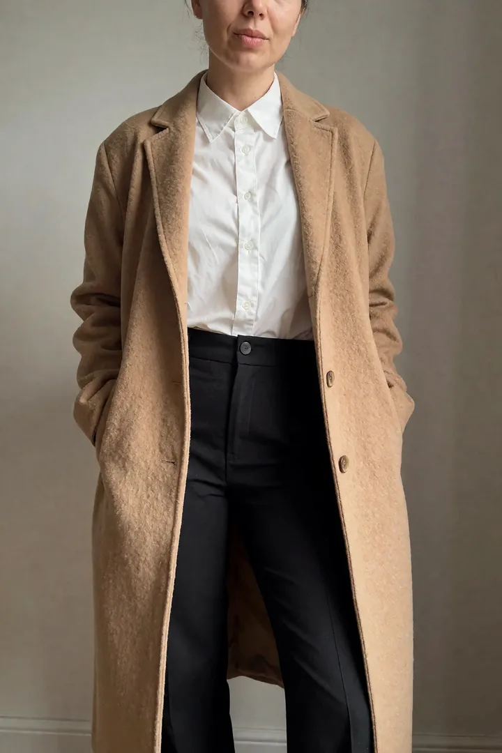 Plano de catálogo: abrigo camel abierto sobre camisa blanca y pantalón negro, fondo liso.